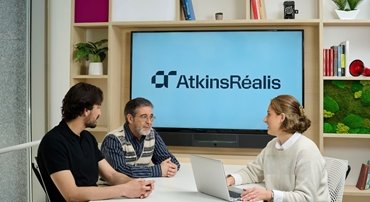 three colleagues talking at a desk wit AtkinsRéalis logo on wall behind them